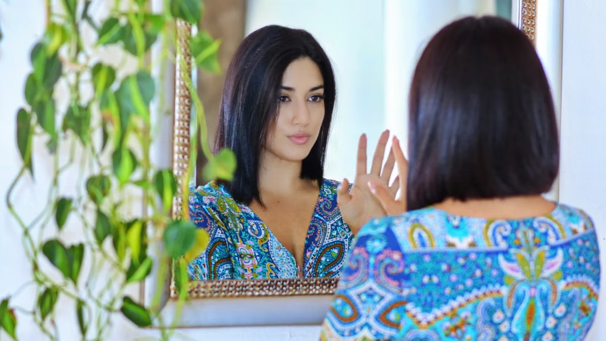 A woman in a vibrant patterned shirt gazes at her reflection in a mirror surrounded by green plants.